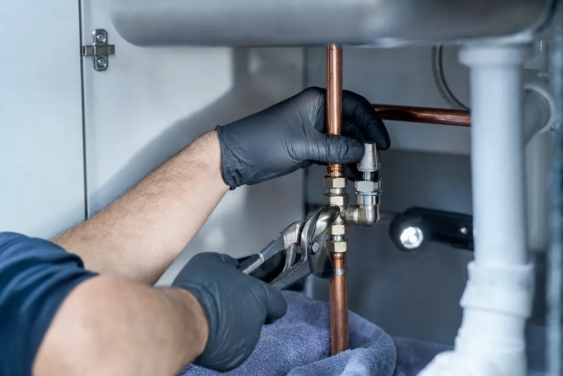 Gloved hands tightening a new shutoff valve on a copper pipe under a kitchen sink.